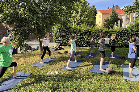 KAT Das KAT-Team beim Yoga im Freien. Personen stehen auf Matten. Im Hintergrund ein Baum und Sträucher.