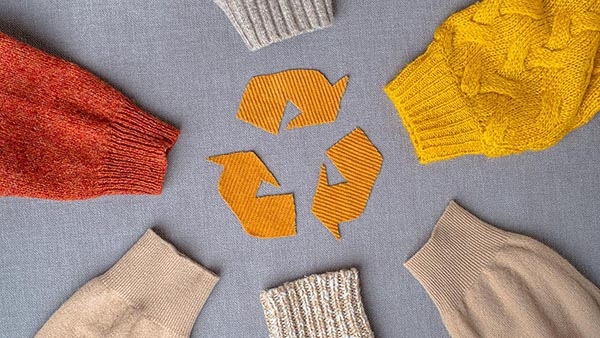 Recycling-Symbol aus Stoffstücken, umgeben von farbigen Strickpullovern auf grauem Hintergrund.