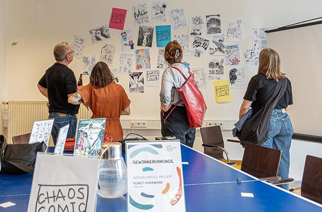 Karl-Heinz Mierke, buehnenlicht Menschen betrachten Comic-Papierzeichnungen an einer Wand; Tisch mit Ausstellungsunterlagen.