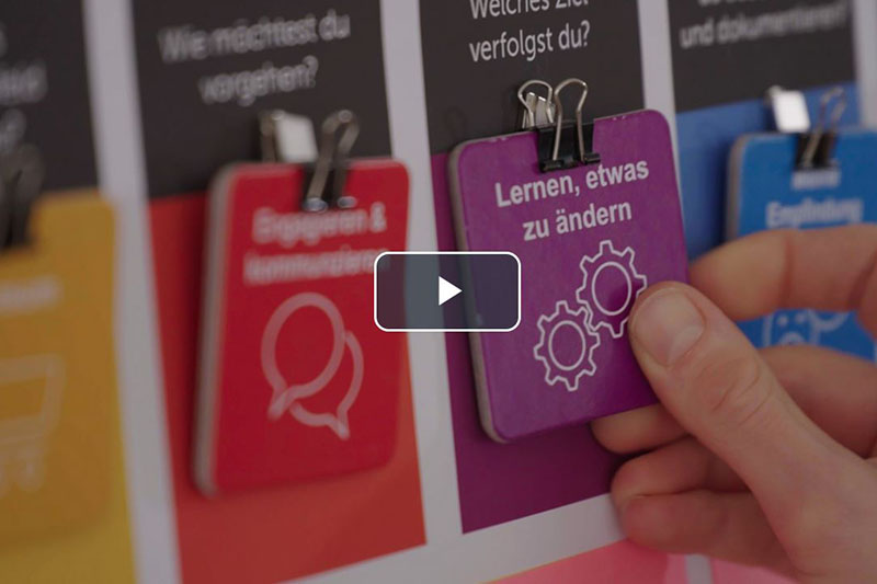 Eine Hand hält lila Karte mit Aufschrift „Lernen, etwas zu ändern“ vor buntem Plakat.