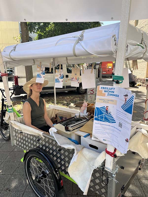 Ein Lastenrad mit einer Person im Wagen steht im Freien vor einem Freiluftwohnzimmer.