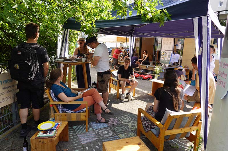 Eine Sitzgruppe im Freien unter einem Zelt, Menschen unterhalten sich, die Sonne scheint. 