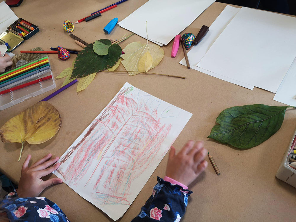 Kind zeichnet mit Buntstiften Blätter auf Papier, um Naturmotive mit Farben darzustellen.