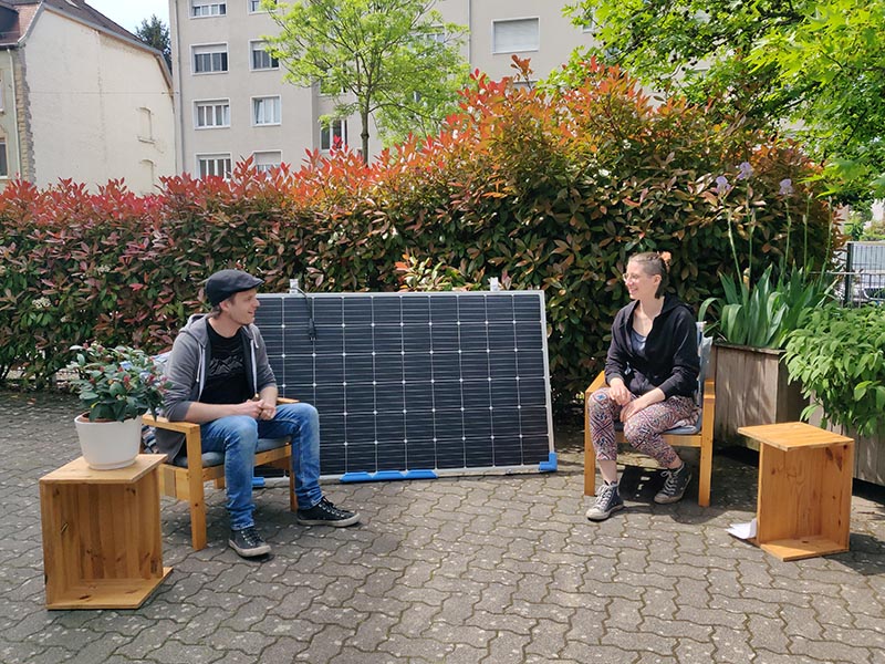 Zwei Personen sitzen im Hof vor einem Solarpanel zwischen Holzstühlen mit Pflanzen.