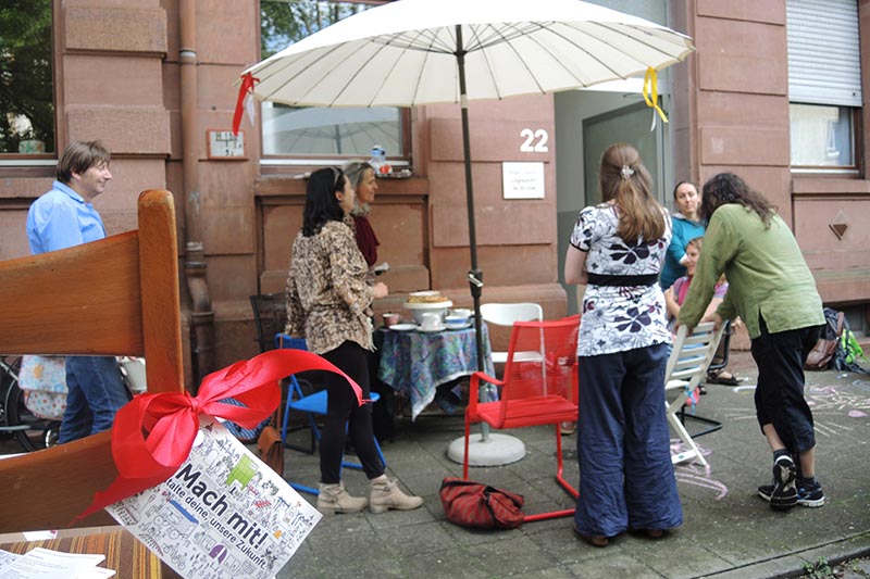 Menschen versammeln sich bei einem Straßenfest mit Sonnenschirm und Tischen vor einem Haus.