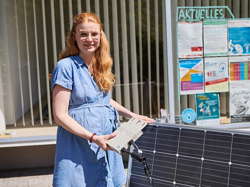 Helena Trenks in blauem Kleid überprüft Solarpanel mit angeschlossenem Wechselrichter.