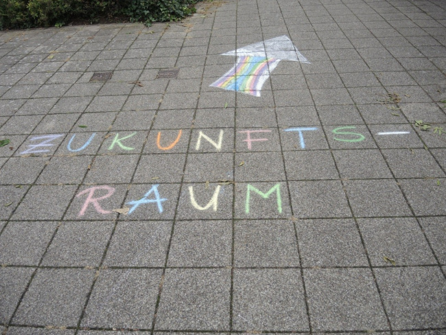 Bunter Kreideschriftzug "Zukunftsraum" mit einem Pfeil auf einem Gehweg aus Pflastersteinen.