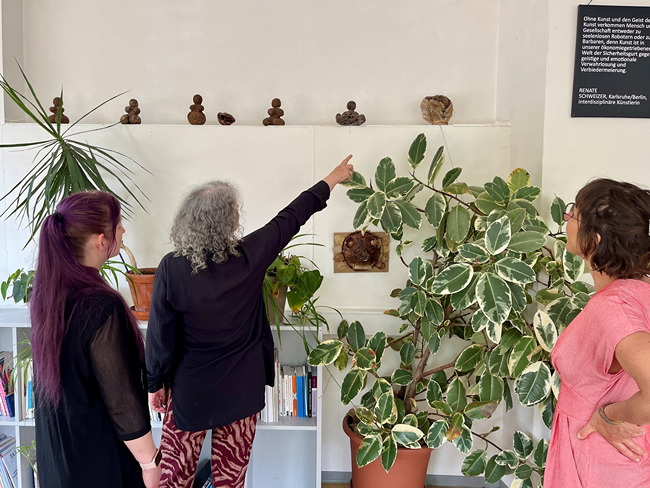 Drei Frauen betrachten kleine Skulpturen an einer wei&szlig;en Wand neben einer gro&szlig;en Zimmerpflanze.