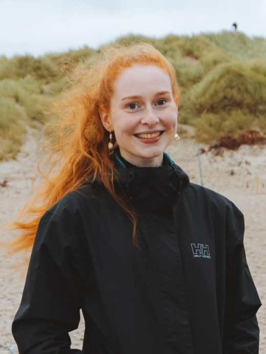 Eine Person am Strand, die roten Haare wehen im Wind.
