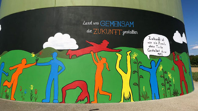 Farbiges Gemälde mit Menschenfiguren und dem Schriftzug „Lasst uns gemeinsam die Zukunft gestalten“ an einem Windradsockel.