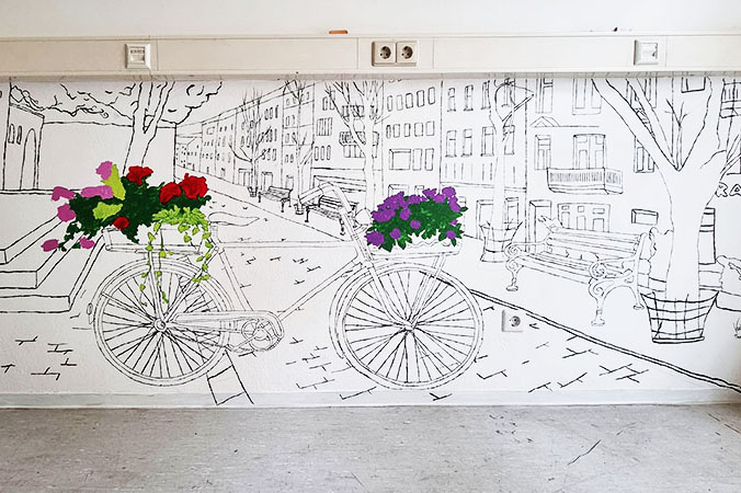 Wandgem&auml;lde einer Stadtstra&szlig;e mit einem Fahrrad, das farbige Blumenk&ouml;rbe tr&auml;gt.