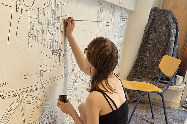 Person zeichnet eine gro&szlig;formatige Stadtszene mit einem Fahrrad an eine wei&szlig;e Wand.