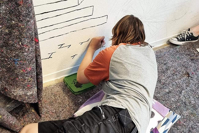 Person zeichnet eine schwarz-wei&szlig;e Grafik an eine Wand.