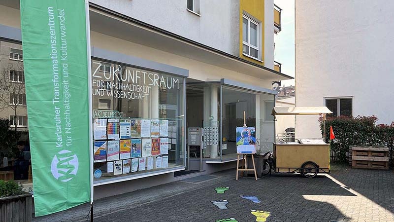 Eingang des Zukunftsraums für Nachhaltigkeit und Wissenschaft am KIT mit Infoplakaten und KAT-Fahne davor.