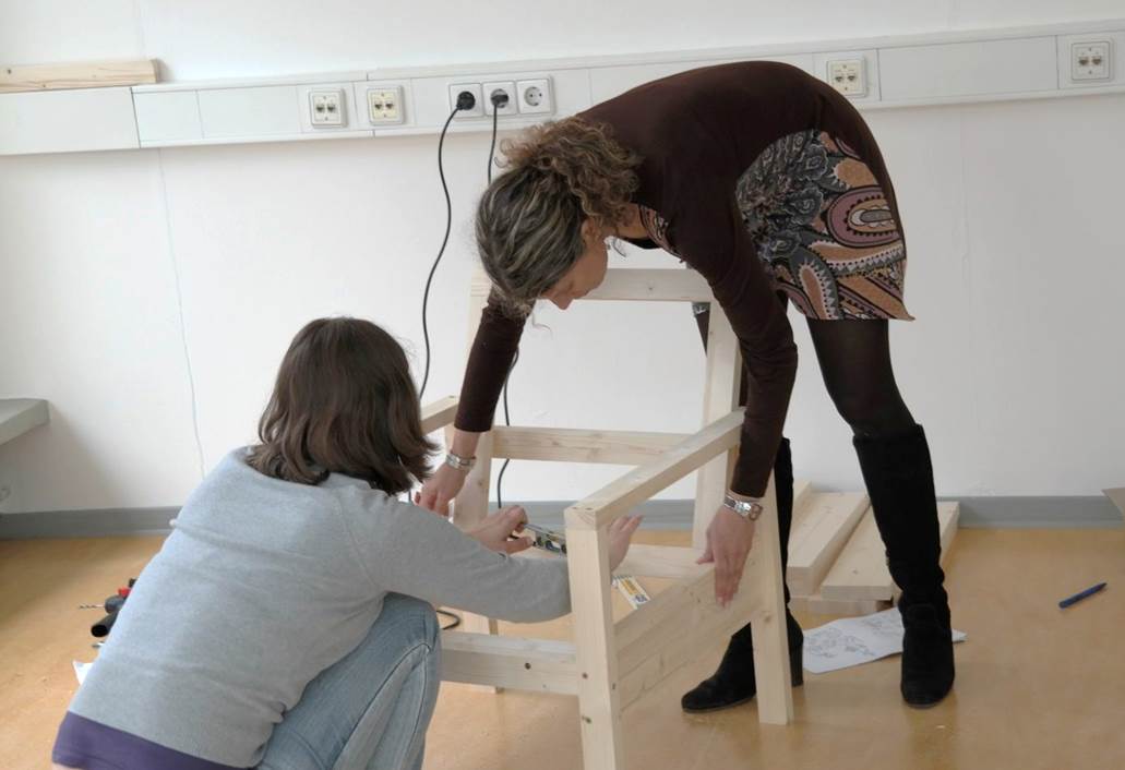 Zwei Frauen bauen gemeinsam einen Holzstuhl in einem Zimmer zusammen.