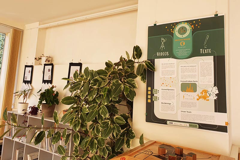 Ein Poster vom Future Fiction-Wettbewerb hängt an der Wand, daneben ein Sideboard mit einer Pflanze und weiteren ausgestellten Exponaten.